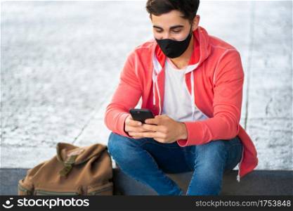 Portrait of young man using his mobile phone while sitting outdoors at the street. Man wearing face mask. Urban concept.