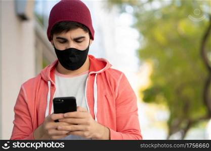 Portrait of young man using his mobile phone while walking outdoors on the street. Man wearing face mask. Urban concept.