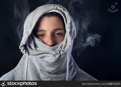portrait of young man wearing a grey hood with half face covered and smoke around him against black background