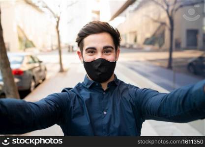 Portrait of young man wearing protective mask and taking a selfie while standing outdoors on the street. New normal lifestyle concept.. Portrait of young man taking a selfie outdoors.