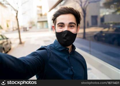 Portrait of young man wearing protective mask and taking a selfie while standing outdoors on the street. New normal lifestyle concept. 