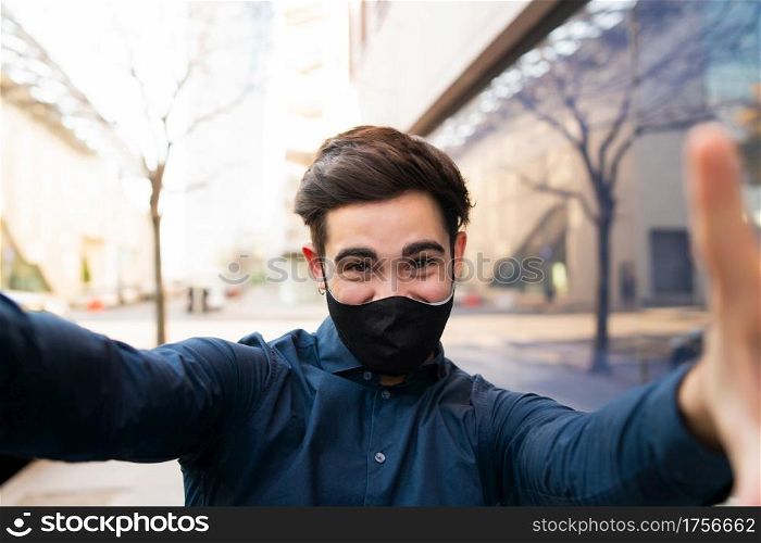 Portrait of young man wearing protective mask and taking a selfie while standing outdoors on the street. New normal lifestyle concept.