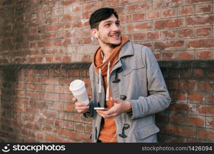 Portrait of young man with earphones and a cup of coffee against brick wall. Outdoors. Urban concept.