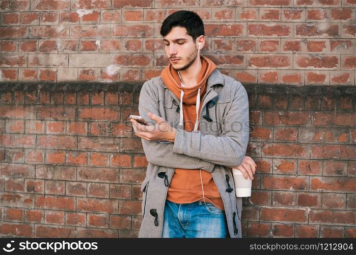 Portrait of young man with headphones and using mobile phone against brick wall. Outdoors. Urban concept.