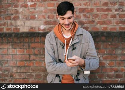 Portrait of young man with headphones and using mobile phone against brick wall. Outdoors. Urban concept.