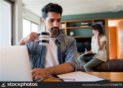 Portrait of young man working with a laptop from home while woman talking on phone at background. Home office concept. New normal lifestyle.