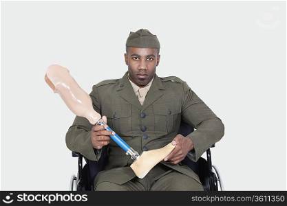 Portrait of young military officer in wheelchair holding prosthesis leg over gray background