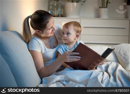 Portrait of young mother lying in bed with her baby at night and holding big book
