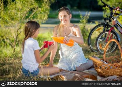 Portrait of young mother pouring juice from bottle into daughter's cup on picnic at park