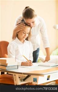 Portrait of young mother praising daughter doing homework at desk