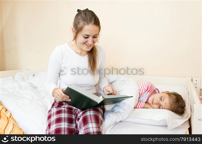 Portrait of young mother sitting on bed and reading bedtime story to daughter
