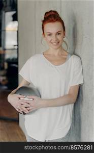 Portrait of young motivated sportive redhaired fitness woman standing in gym with small exercise ball, leaning on concrete wall and looking happily into camera, wearing white t-shirt and grey leggings. Cute young red haired woman with pilates ball relaxing after workout in fitness studio