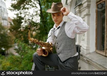 Portrait of young musician wearing heat and stylish outfit holding saxophone musical instrument. Handsome jazzman looking at camera. Portrait of young musician holding saxophone musical instrument