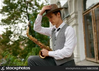 Portrait of young musician wearing heat and stylish outfit holding saxophone musical instrument. Handsome jazzman looking at camera. Portrait of young musician holding saxophone musical instrument