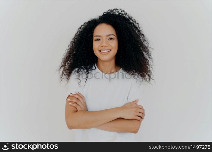 Portrait of young pleasant looking woman with curly hair, keeps hands crossed, wears white t shirt, smiles broadly at camera, has timid shy expression, poses indoor, has natural beauty. Emotions