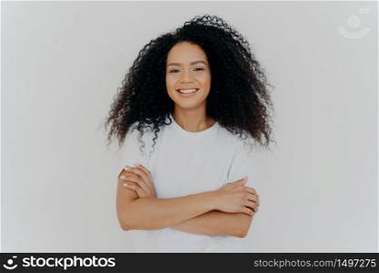 Portrait of young pleasant looking woman with curly hair, keeps hands crossed, wears white t shirt, smiles broadly at camera, has timid shy expression, poses indoor, has natural beauty. Emotions