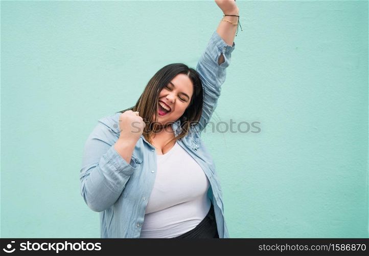 Portrait of young plus size woman celebrating victory outdoors against light blue background. Success concept.