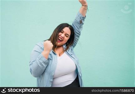 Portrait of young plus size woman celebrating victory outdoors against light blue background. Success concept.