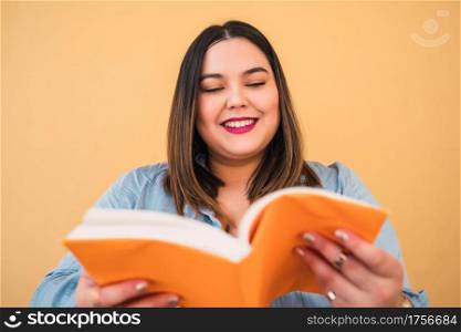 Portrait of young plus size woman enjoying free time and reading a book while standing against yellow background. Lifestyle concept.