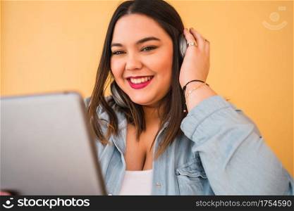 Portrait of young plus size woman listening to music with headphones and digital tablet outdoors against yellow background.