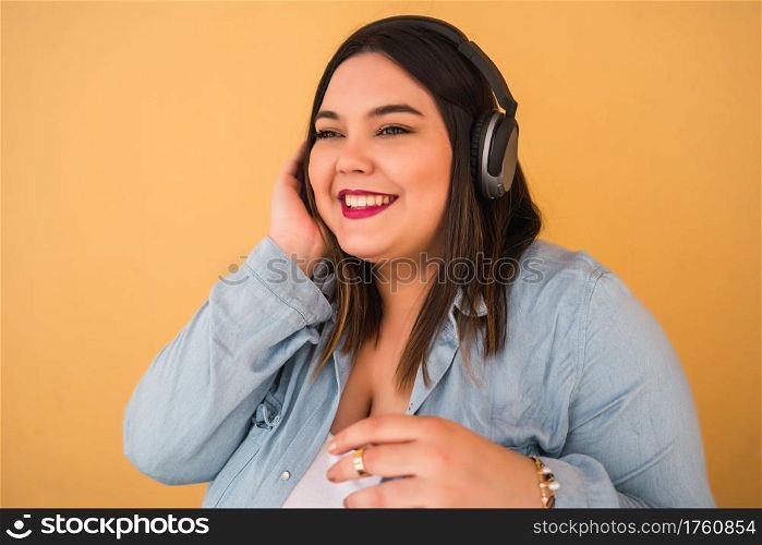 Portrait of young plus size woman listening to music with headphones outdoors against yellow background.