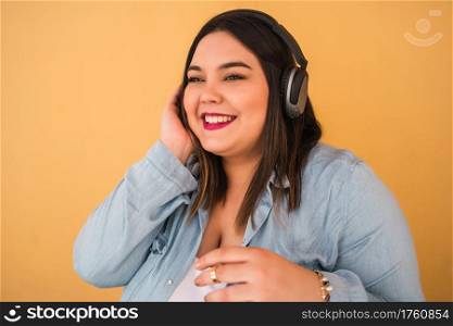 Portrait of young plus size woman listening to music with headphones outdoors against yellow background.