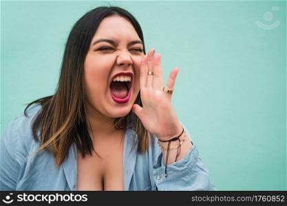 Portrait of young plus size woman shouting and screaming to someone while standing against light blue wall outdoors.