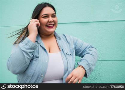 Portrait of young plus size woman smiling while talking on the phone outdoors against light blue background. Communication concept.