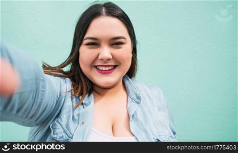 Portrait of young plus size woman taking a selfie while standing against light blue wall, outdoors.
