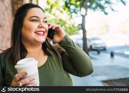 Portrait of young plus size woman talking on the phone while holding a cup of coffee outdoors in the street. Urban concept.