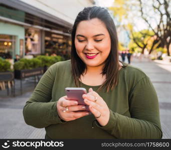 Portrait of young plus size woman typing text message on her mobile phone outdoors on the street. Technology concept.