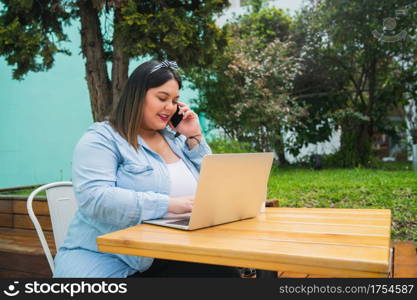 Portrait of young pluse size woman using her laptop and talking on the phone while sitting outdoors at coffee shop.