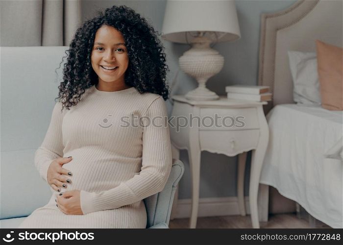 Portrait of young positive pregnant woman sitting in bedroom and looking at camera with bright smile, holding her belly and enjoying pregnancy time while relaxing on armchair at home. Young happy pregnant woman with curly hair holding her belly and smiling at camera