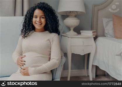 Portrait of young positive pregnant woman sitting in bedroom and looking at camera with bright smile, holding her belly and enjoying pregnancy time while relaxing on armchair at home. Young happy pregnant woman with curly hair holding her belly and smiling at camera
