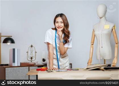 Portrait of young pretty female fashion designer stylish standing and hand on shin with smile feeling proud her business during working at fashion studio, full of tailoring tools and equipment