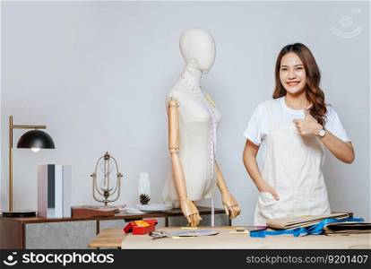 Portrait of young pretty female fashion designer stylish standing and raise hand thump up with smile feeling proud her business during working at fashion studio, full of tailoring tools and equipment