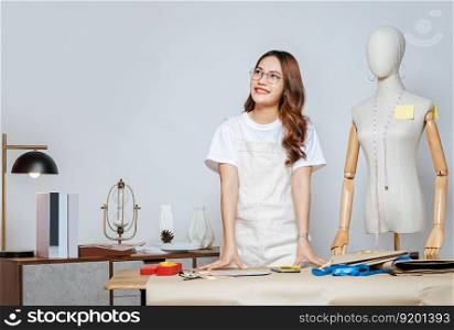 Portrait of young pretty female fashion designer stylish wearing eyeglasses standing and smile with proud during working at fashion studio, full of tailoring tools and equipment