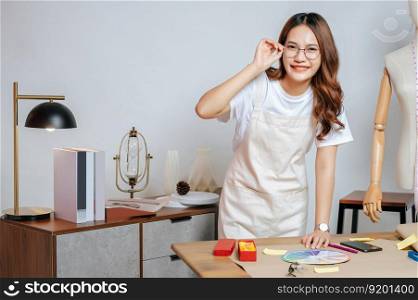 Portrait of young pretty female fashion designer stylish wearing eyeglasses standing and smile with proud during working at fashion studio, full of tailoring tools and equipment