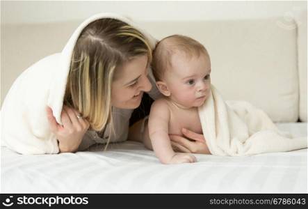 Portrait of young smiling mother lying on bed with her baby son under white blanket