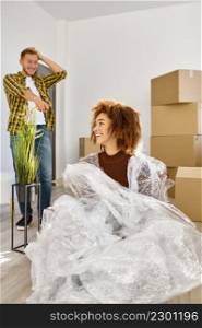 Portrait of young smiling woman wrapped in packing bubble tape fooling around during home moving. Woman wrapped in packing bubble tape portrait