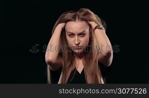 Portrait of young stressed woman pondering over problem with her hands on head against black background. Pensive attractive girl with long blonde hair and blue eyes thinking hard to find solution and looking at camera with dissapointed gaze.