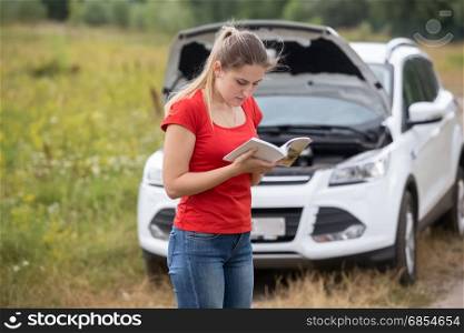 Portrait of young stressed woman standing at broken car and reading owner manual