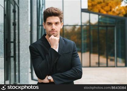 Portrait of young successful pensive businessman in city. Man in business jacket on office building background. Handsome person ponders about work.. Portrait of young successful pensive businessman in city. Man in business jacket on office building background. Handsome person ponders about work