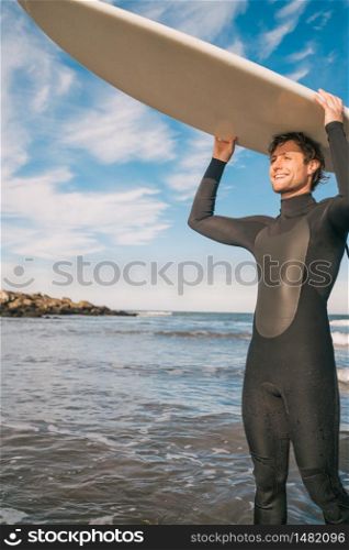 Portrait of young surfer at the beach holding up his surfboard and wearing a black surfing suit. Sport and water sport concept.