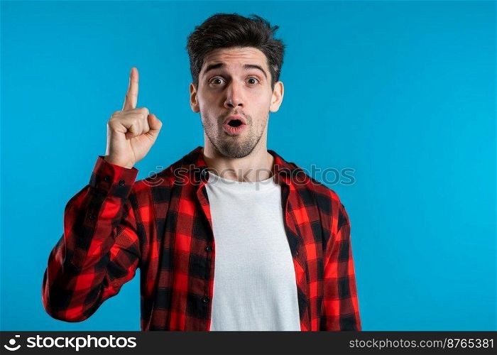 Portrait of young thinking pondering man in red having idea moment pointing finger up on blue studio background. Smiling happy student guy showing eureka gesture. Portrait of young thinking man in red having idea moment pointing finger up