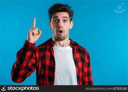 Portrait of young thinking pondering man in red having idea moment pointing finger up on blue studio background. Smiling happy student guy showing eureka gesture. Portrait of young thinking man in red having idea moment pointing finger up
