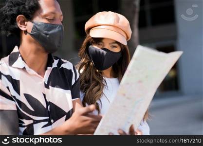 Portrait of young tourist couple using protective mask and looking at map while looking for directions outdoors. Tourism concept. New normal lifestyle concept.