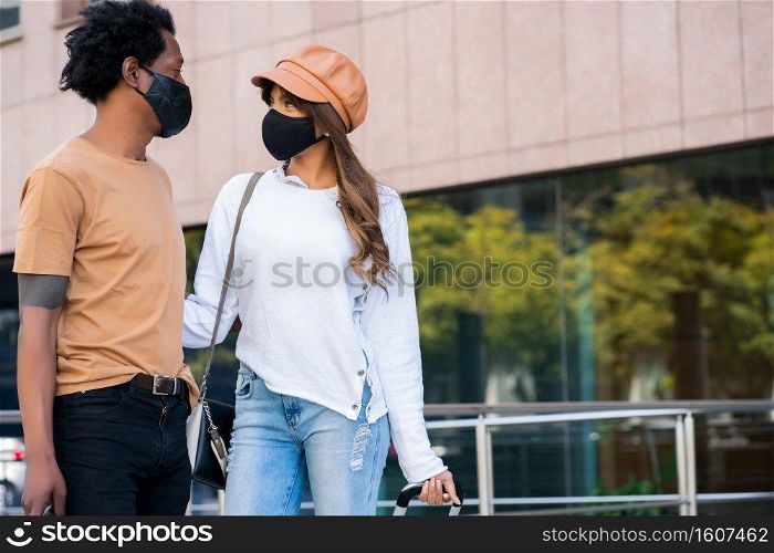 Portrait of young tourist couple wearing protective mask and carrying suitcase while walking outdoors on the street. Tourism concept. New normal lifestyle concept.