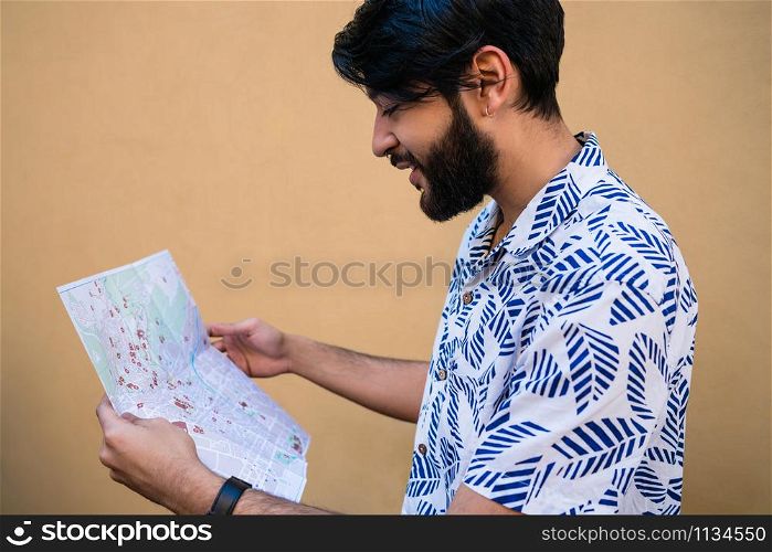 Portrait of young tourist man wearing summer clothes and holding a map looking for directions against yellow background. Holiday and travel concept.