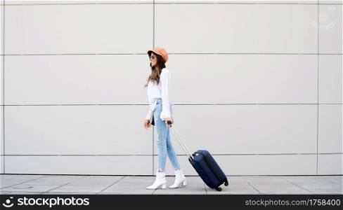 Portrait of young traveler woman carrying a suitcase while walking outdoors on the street. Tourism concept.